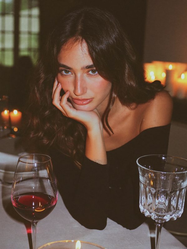 Illustration: a brunette in an off-shoulder black top, chin in her hand, at a candlelit dinner table with wine glasses