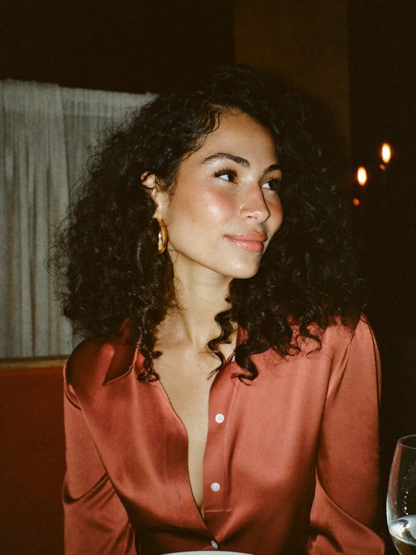 Illustration: a woman with dark curly hair and gold hoops, in a rust satin shirt, smiling at a restaurant table at night