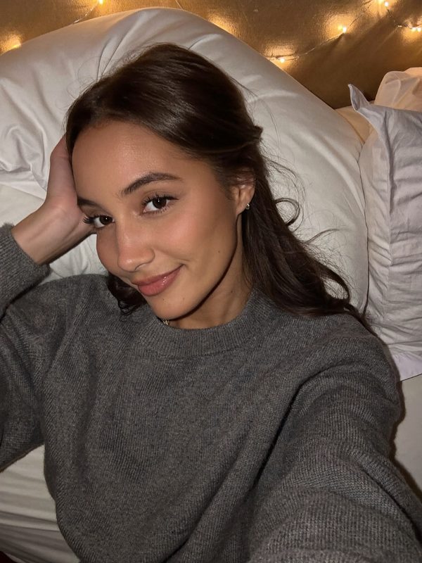 Illustration: a smiling brunette in a grey sweater taking a selfie in bed, fairy lights behind her